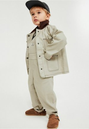 Junge im beigen Jackett und Hose mit braunem Kragen und Schuhen, trägt eine schwarze Mütze, steht mit der Hand in der Tasche und schaut nach oben und links.