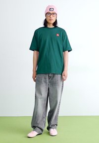 Tricou verde din bumbac cu un mic logo roșu, asociat cu blugi denim de un gri deschis și adidași roz. Fundalul este alb simplu.