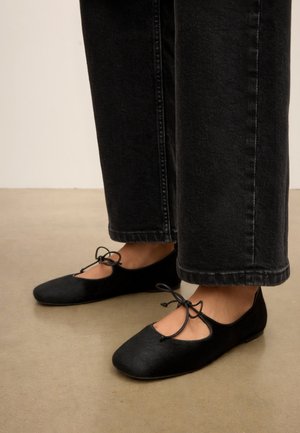 Voeten met zwarte ballerina's met enkelbandjes en strikjes, gecombineerd met zwarte wijde broekspijpen jeans, staand op een beige vloer.