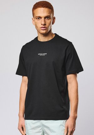 Junger Mann mit kurzen roten Haaren, der ein schwarzes T-Shirt mit der Aufschrift "Weekend Offender Admit Nothing" und helle Hosen vor einem einfarbigen Hintergrund trägt.