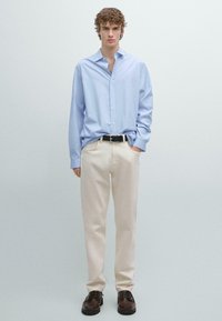 Světle modrá košile na knoflíky s dlouhými rukávy, béžové kalhoty a tmavě hnědé boty. Košile má uvolněný střih a měkkou texturu.