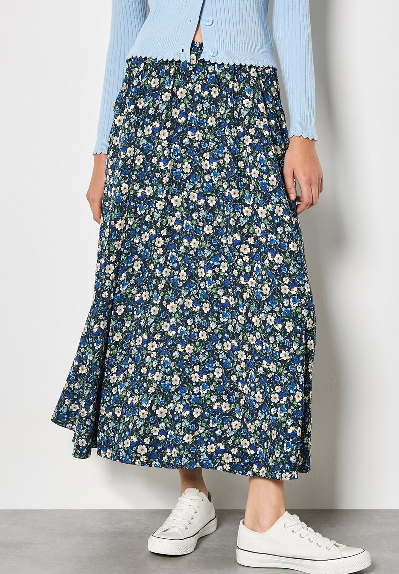Femme portant une jupe midi bleue à fleurs, un cardigan bleu clair boutonné et des baskets blanches, debout sur un sol gris contre un mur blanc.