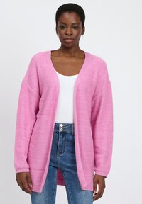 Roze gebreide cardigan met een losse pasvorm, lange mouwen en een V-hals. Heeft subtiele sprankelingen en geribbelde manchetten, gecombineerd met een witte top en denim jeans.
