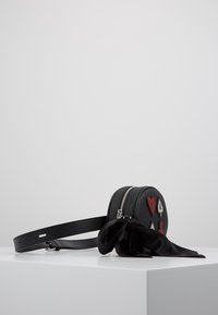 Svart rund läderbältesväska med lappar av spelkortssymboler och en knuten svart satin scarf på ett vitt podium.