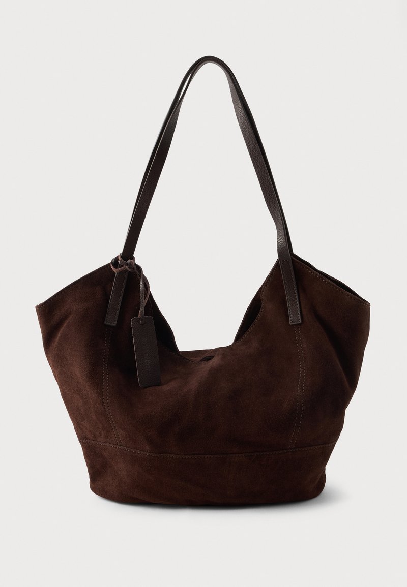 Bolso de ante marrón con una forma suave y caída. Presenta asas de cuero y un acento de etiqueta. Textura lisa con costuras curvas.