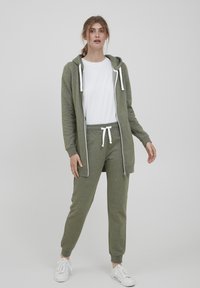 Olijfgroene hoodie met trekkoorden, bijpassende joggingbroek met elastische tailleband, witte t-shirt eronder, en witte sneakers. Zachte, gestructureerde stof.