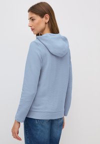 Felpa leggera blu con superficie texturizzata, dotata di cappuccio e maniche lunghe. Indossata con jeans di denim, che mostrano un taglio e un design casual.