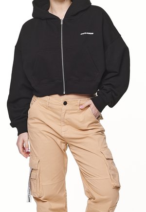 Femme portant un sweat à capuche court noir zippé avec poches avant et pantalon cargo beige avec poches latérales, main reposant dans la poche.