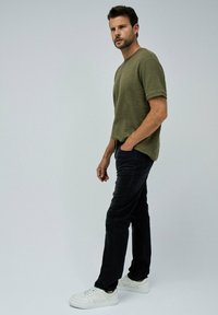 Camisa de manga corta verde oliva con rayas texturizadas, combinada con jeans negros y zapatillas blancas, mostrando un atuendo casual.