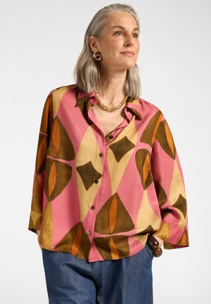 Femme aux cheveux gris portant un chemisier coloré à motifs rose, orange et marron, un jean bleu, un collier en or et de grandes boucles d'oreilles rondes.