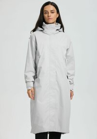 Mujer con un abrigo de lluvia largo y gris claro con capucha y mangas largas, de pie frente a un fondo claro y sencillo.