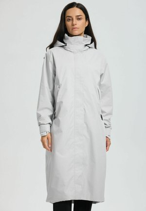 Kvinna som bär en lång, ljusgrå huva regnjacka med långa ärmar, står mot en enfärgad ljus bakgrund.