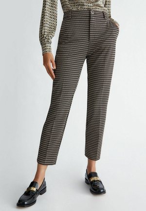 Femme portant un pantalon à motifs longueur cheville, une blouse assortie à motifs et des mocassins noirs avec des détails dorés, debout les mains dans les poches.