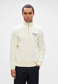 Vyras vilkintis kreminiu ketvirčio užtrauktuko džemperiu su užrašu "Gant 1949 American Sportswear", derinamas su tamsiais džinsais, stovi prieš paprastą foną.