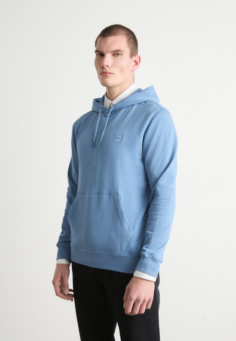 Sweatshirt azul claro com capuz, bolso frontal e punhos elásticos; apresenta um pequeno patch de logo no peito. Textura de tecido macio.