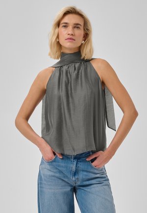 Lyshåret kvinde med hænderne i lommerne iført en ærmeløs grå højhalset top og blå jeans mod en ensfarvet lys baggrund.