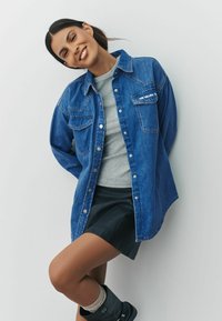 Denim shirt in blue with two chest pockets, button closure, and subtle branding. Worn over a grey top and paired with a black leather skirt.