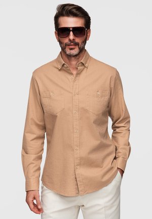 Homme portant une chemise beige boutonnée avec poches poitrine et un pantalon blanc, posant avec une main dans la poche et des lunettes de soleil foncées.