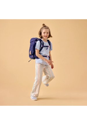 Kind mit kurzen Haaren, der ein hellblaues Hemd, gestreifte Hosen und Turnschuhe trägt und einen dunkelblauen Rucksack trägt, während es ein Bein anhebt.