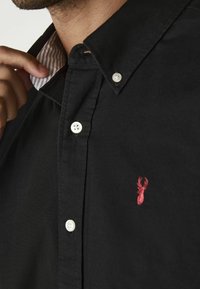 Mann, der den Kragen eines schwarzen Hemdes mit weißen Knöpfen und einem roten gestickten Hirschlogo auf der Brust anpasst.