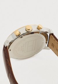 Πίσω μέρος ενός ρολογιού χειρός Tommy Hilfiger με θήκη από ανοξείδωτο ατσάλι, κουμπιά σε χρυσό τόνο και καφέ δερμάτινο λουράκι.