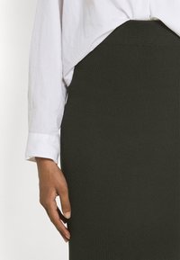 Ribbad mörkgrön pennkjol i stickad tyg, parat med en vit skjorta. Har en figurnära silhuett och medelhög midja.