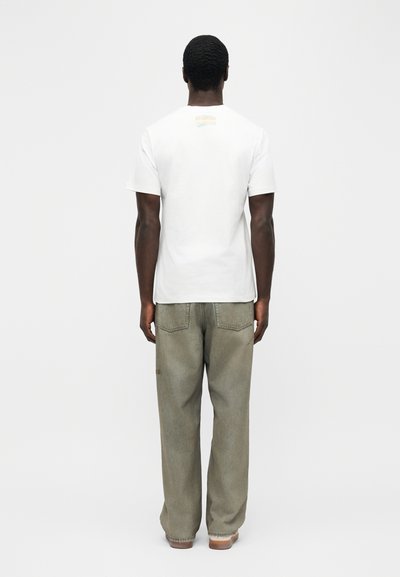 Άνδρας με την πλάτη, φοράει απλό λευκό κοντομάνικο πουκάμισο και φαρδιά ξεβαμμένα γκρι τζιν, με φόντο λευκό.