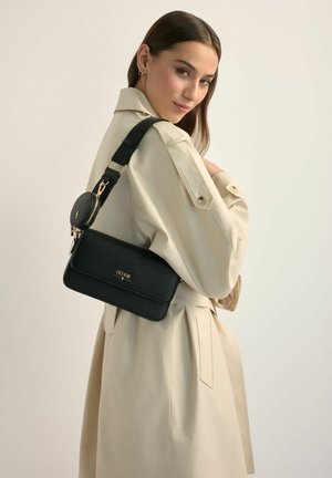 Młoda kobieta w beżowym płaszczu trencz, niosąca czarną torbę na ramię z małą okrągłą sakiewką na pasku, spogląda przez ramię za siebie.