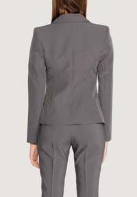 Blazer grigio aderente, con spalle strutturate e colletto a punta. Tessuto liscio con silhouette sartoriale, abbinato a pantaloni coordinati.