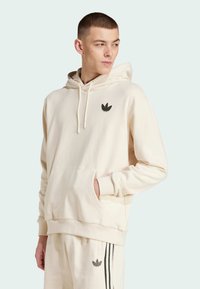 Beige hooded sweatshirt made of soft fabric with a front pocket. Features a black Adidas logo on the chest and ribbed cuffs at the sleeves.