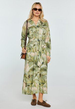 Kobieta w długiej zielonej sukience w kwiaty zapinanej na guziki z paskiem, brązowych mokasynach, brązowej torebce na ramię i ciemnych okularach przeciwsłonecznych, stojąca na jednolitym tle.