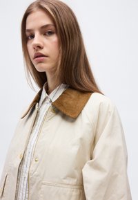 Νεαρή γυναίκα με μακριά ανοιχτοκάστανα μαλλιά, που φοράει μπεζ σακάκι με καφέ γιακά από κοτλέ και ένα λευκό πουκάμισο από κάτω.