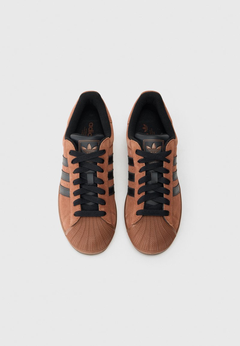 Paar braune Adidas-Sneaker aus Wildleder mit schwarzen Streifen, schwarzen Schnürsenkeln und Zehenkappen im Shell-Stil, von oben auf weißem Hintergrund gesehen.
