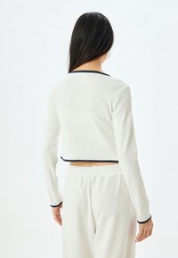 Witte geribbelde crop top met lange mouwen, met een ronde halslijn met marineblauwe afwerking. Gecombineerd met crèmekleurige broek met elastische taille.