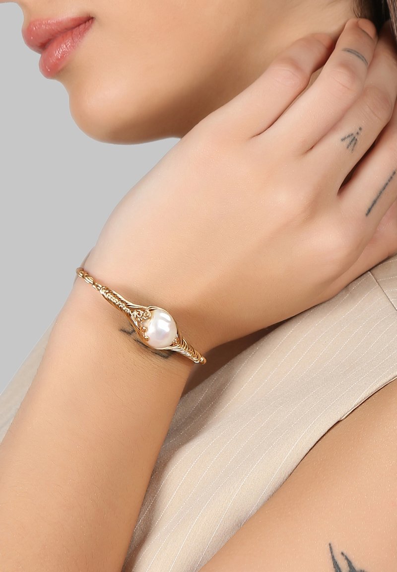 Gold bracelet featuring a prominent white pearl, with intricate twisted design and engraved accents, worn on a hand with visible tattoos.