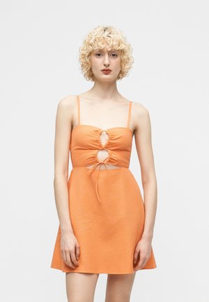 Νεαρό άτομο με σγουρά ξανθά μαλλιά, ντυμένο με πορτοκαλί αμάνικο φόρεμα με ανοίγματα και δέσιμο μπροστά, που στέκεται μπροστά σε απλό φόντο.