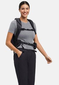 Czarny plecak z regulowanymi paskami o smukłym designie. Osoba ma na sobie szarą koszulę w paski i czarne spodnie, ręce w kieszeniach.