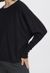 Pull noir ample avec un col rond et des épaules tombantes. Texture douce, manches longues et poignets côtelés.