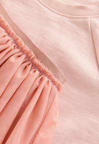 Vêtement rose clair avec une partie supérieure lisse et un overlay en tulle texturé froncé en bas ; détail de taille élastiquée.