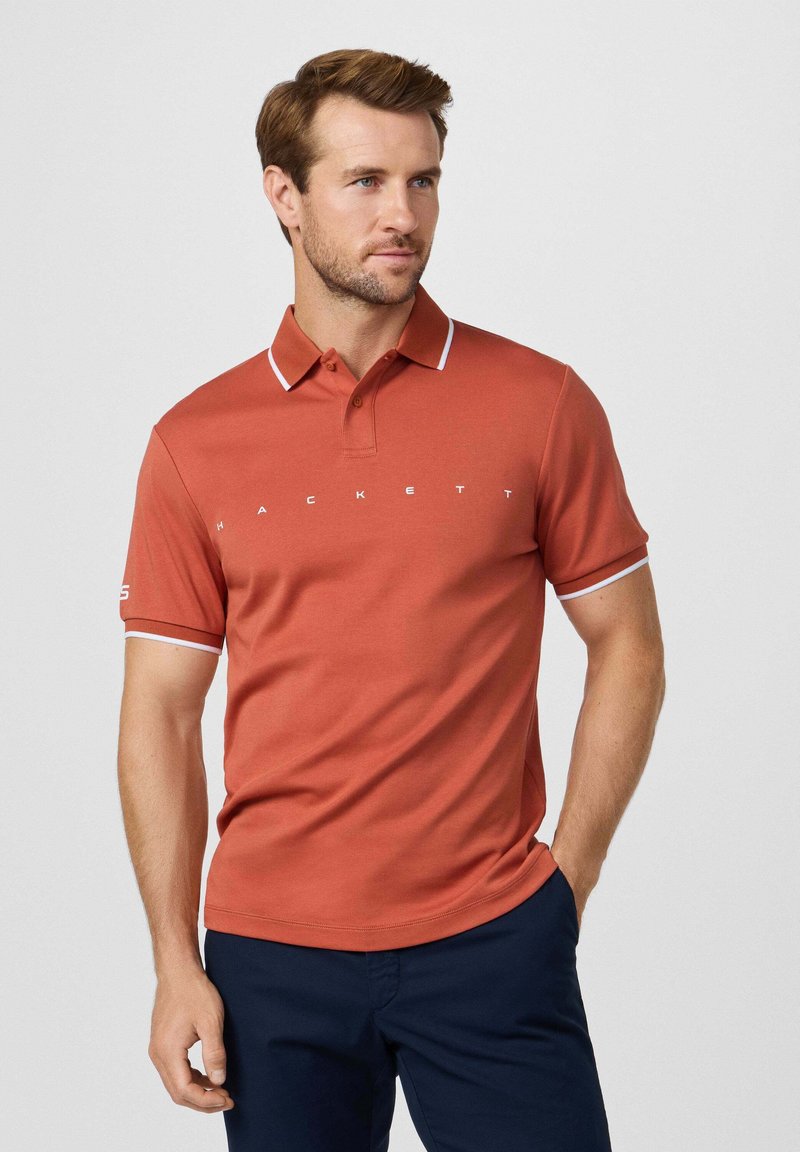 Polo color ruggine con dettagli bianchi su colletto e maniche; presenta testo del logo bianco sul petto. Maniche corte e vestibilità classica.