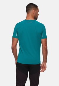 Man som bär en teal kortärmad träningsskjorta med Mammut-logotyp på övre delen av ryggen och svarta byxor, sedd bakifrån mot vit bakgrund.