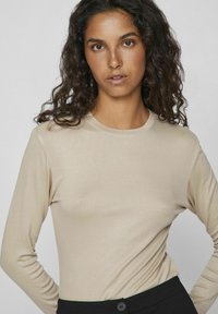 Beige langermet topp laget av glatt, elastisk stoff, med rund hals og tettsittende silhuett, kombinert med svarte bukser.