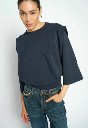 Vrouw draagt een donkerblauw losvallend shirt met mouwen en blauwe spijkerbroek met een kettingdetail op de zak, staand tegen een effen lichte achtergrond.