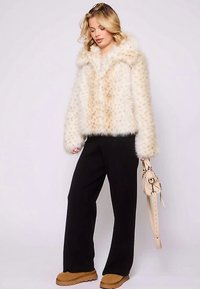 Kort jakke i imiteret pels i cremefarve med en fluffy tekstur, kombineret med sorte bukser med vide ben og brune støvler, med en lys håndtaske i hånden.