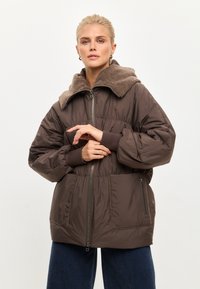 Bruine pufferjack met een getextureerde taupekleurige capuchon, een ritssluiting aan de voorkant, elastische manchetten en twee voorzakken. Gemaakt van een glanzend, waterafstotend materiaal.