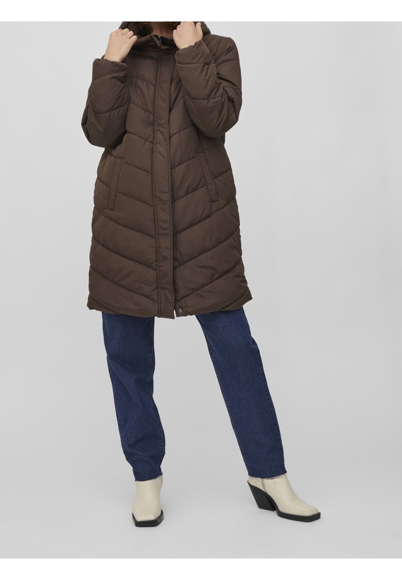 Femme portant un long manteau matelassé marron, un jean bleu et des bottines blanches à talons, debout devant un fond clair uni.
