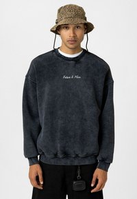 Donkergrijze sweatshirt van zacht materiaal, met 'Future Is Here' geborduurd in het wit. Gecombineerd met een lederen bucket hat met luipaardprint en zwarte broek.