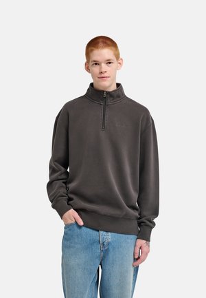 Junger Mann mit kurzen roten Haaren, der einen dunkelgrauen Sweatshirt mit Reißverschluss und hellblaue Jeans trägt, steht mit einer Hand in der Tasche.