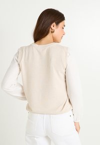 Suéter beige con un frente de punto texturizado y mangas largas de canalé blancas, combinado con jeans blancos. La vista trasera resalta el contraste de colores.
