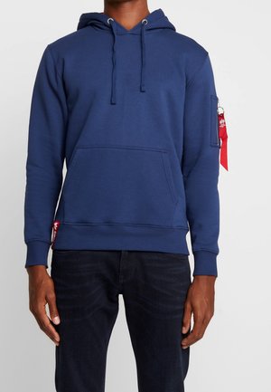 Mężczyzna w granatowej bluzie z kapturem z kieszenią z przodu i czerwonym suwakiem na lewym rękawie, w ciemnych dżinsach, stojący na białym tle.